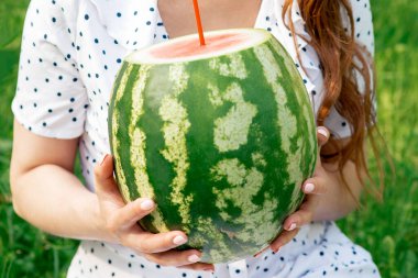 Dışarıdaki çimlerin üzerinde duran kadının ellerinde kokteyl pipetli bir karpuz..