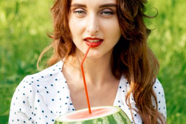 Genç bir kadın karpuz tutuyor dudaklarının yanında kokteyl kamışı dışarıda çimenlerde oturuyor..