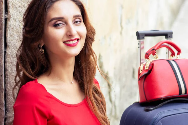 Portrait of beautiful smiling woman with suitcase in red dress over old wall.