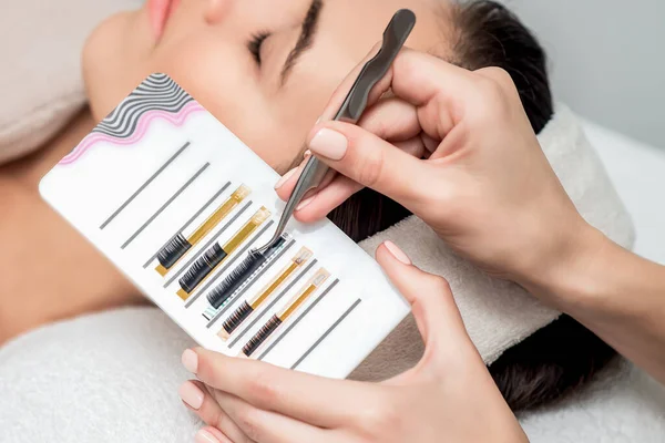 Hands of beautician takes artificial eyelashes by tweezers from tray on background of young woman.