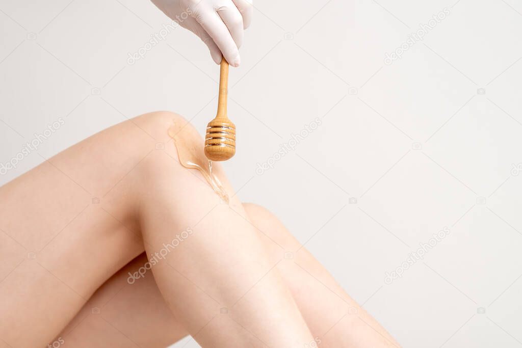 Wax ax flows down the honey stick on leg during hand holding stick on white background. Concept of depilation and epilation.