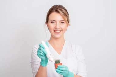 Kozmetik uzmanı kahverengi bir tencerede yeşil kaktüs tıraş ediyor ve beyaz arka planda gülümseyen eldivenler giyiyor. Tüy dökümü, epilasyon ve tüylerin alınması kavramı.