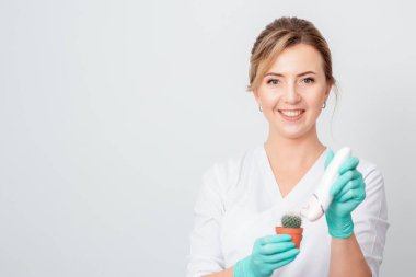 Kozmetik uzmanı kahverengi bir tencerede yeşil kaktüs tıraş ediyor ve beyaz arka planda gülümseyen eldivenler giyiyor. Tüy dökümü, epilasyon ve tüylerin alınması kavramı.