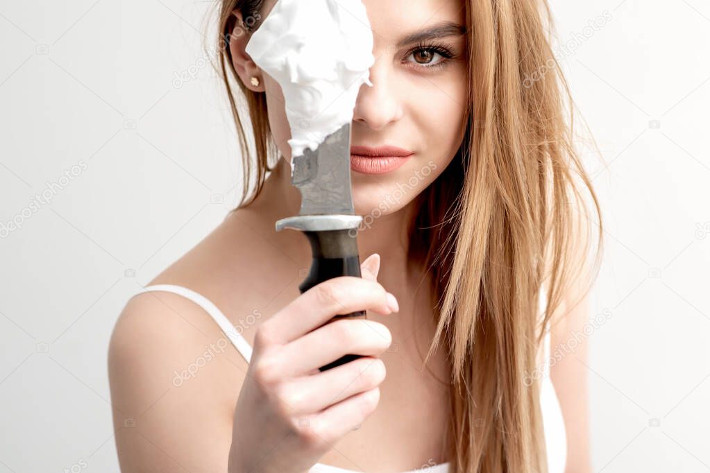 Beautiful young woman is covering her eye by knife with shaving foam on white background.