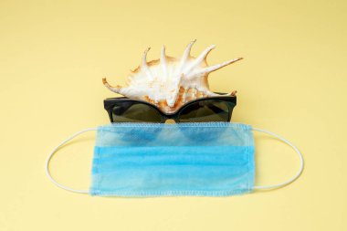 Sarı arka planda güneş gözlüğü ve tıbbi maske takan deniz kabuğu. Seyahat konsepti sırasında Coronavirus veya COVID-19.