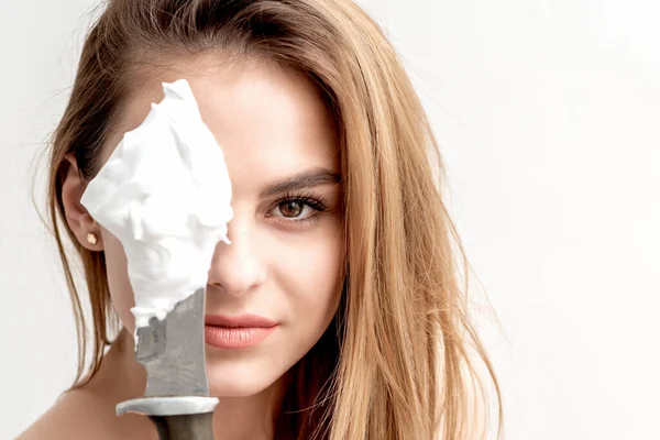 Close-up of knife with shaving foam covering eye of beautiful young woman on white background with copy space.