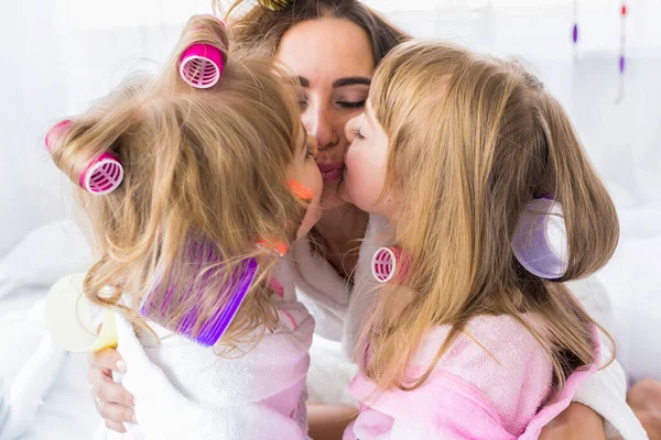 Mother and children are kissing with curlers in hair on the bed in the bedroom. Stay home and family concept.