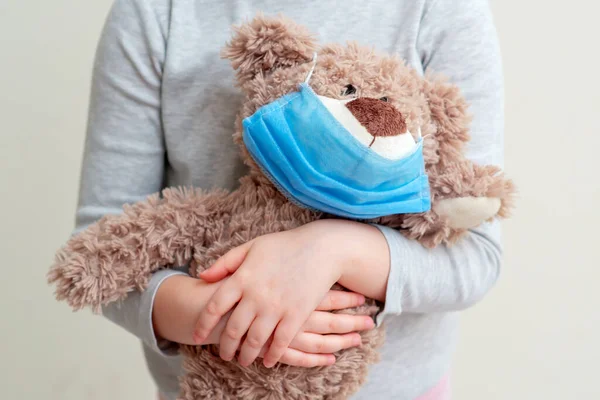 Soft toy bear with protective medical mask in child hands on white background. Health care and virus protection concept.