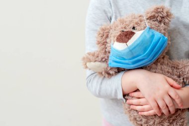 Koruyucu tıbbi maskeli yumuşak oyuncak ayı beyaz arka planda çocuk ellerinde. Sağlık ve virüs koruma kavramı.