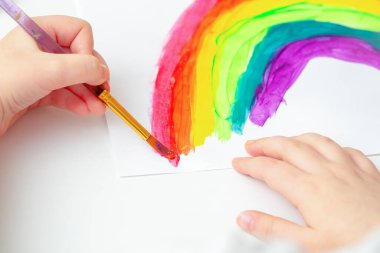 Çocuğun elleri beyaz kağıt yaprağına suluboya ile renkli gökkuşağı çiziyor. Çocukların yaratıcılık kavramı.