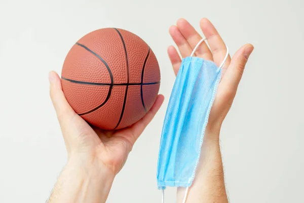 Man hands holding basketball ball with medical protective mask on light background during coronavirus. Basketball paused because coronavirus.