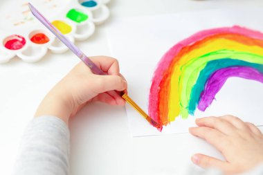 Beyaz kağıda fırça ve boya çizilmiş bir gökkuşağı çizen çocuk ellerinin üst görüntüsü.