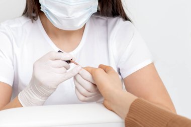 Manikür ustasının elleri manikür salonunda koruyucu maske takan genç bir kadının tırnaklarına oje sürüyor..