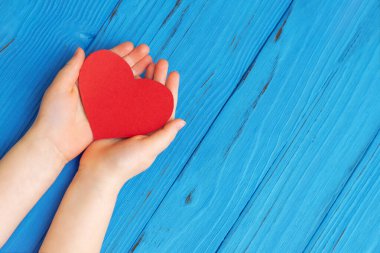 Çocuğun elleri ahşap bir arka planda kırmızı kalbi tutuyor. Sağlık, aşk ve pediatrik kardiyoloji konsepti. Boş boşluk.