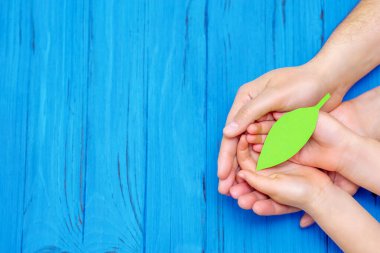 Yeşil kağıt yaprağın üst görüntüsü, ahşap mavi arka planda yetişkin ve çocuğun ellerinde. Dünya Çevre Günü, Dünya Sağlık Günü ve Dünya Günü kavramı. Boşluğu kopyala.