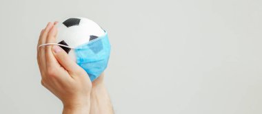 Fotokopi alanı olan gri bir arka planda tıbbi maskeli futbol topu tutan adamın elleri. Karantinaya alınmış futbol.
