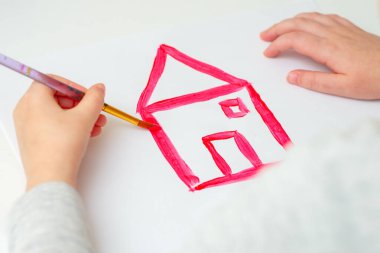 Çocuğun elleri beyaz kağıt üzerinde fırçayla kırmızı ev çiziyor. Sanat eseri konsepti.