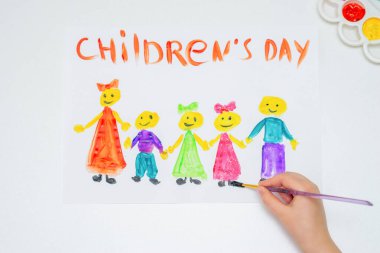 Çocuk elinin en üstteki görüntüsü suluboya fırçayla renkli çocukları çiziyor ve beyaz bir kağıtta Çocuk Günü yazıyor..