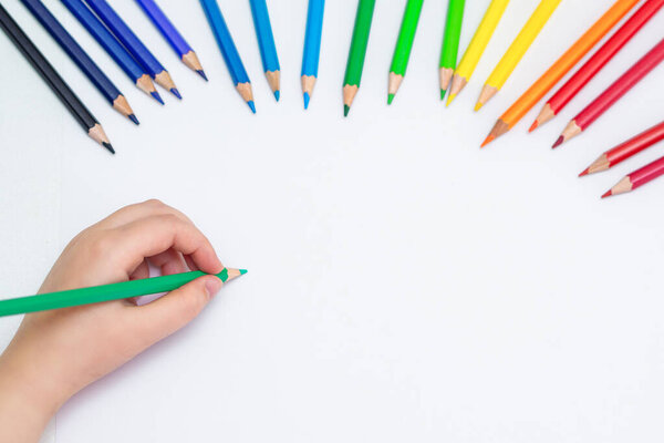 Верхний вид руки ребенка, держащего карандаш зеленого цвета для набросков и рисования с набором цветных карандашей на пустой бумаге. Копирование текста
.