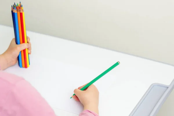 Little girl is drawing by green pencil on white sheet of paper holding bunch of colored pencils in her hand. Selective focus.