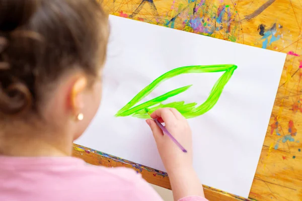 小さな女の子は 白い紙にイーゼルに水彩で筆で緑の葉を描いています 地球の日のコンセプト — ストック写真