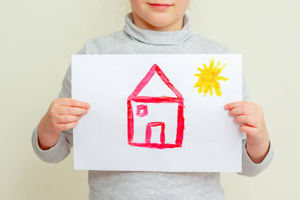 Closeup of Hands of child holding picture of red house with sun covering her face on yellow background. The child learns to draw.