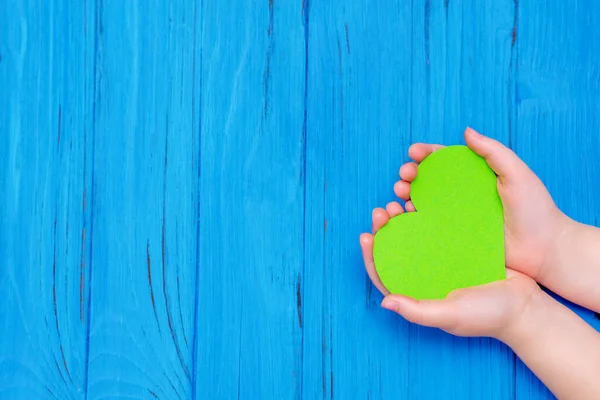 Top view of green heart in hands of child on wooden blue background. Copy space. Concept of environmental protection.