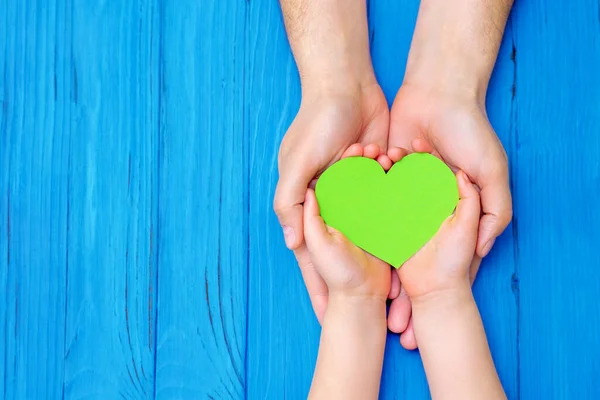 Top view of green heart in hands of adult and child on wooden blue background. Concept of world environment day, world health day and Earth day. Copy space.