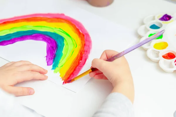 Close up of hand of kid with painted rainbow on white paper during the Covid-19 quarantine at home. Children\'s creativity.