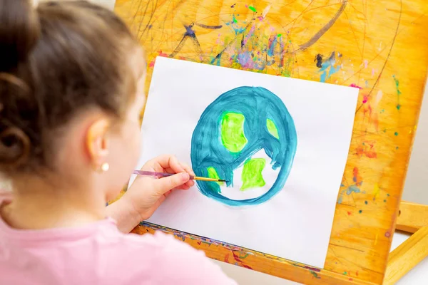 Little girl is drawing earth by watercolors on white paper on a wooden easel. Earth Day concept.