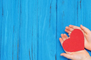 Mavi ahşap arka planda kırmızı bir kalbi tutan bebek elleri. Metin için boş boşluk.