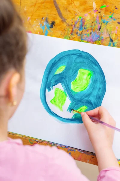 Close up of hand of kid drawing a picture of earth globe. Child is painting Earth with brush by acrylic color on white paper on an easel. Earth day concept.