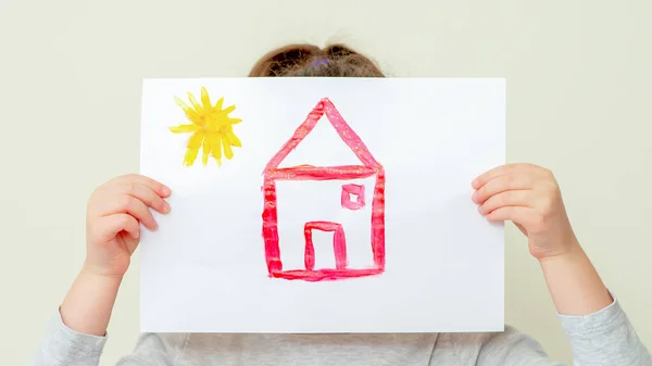 Hands of child holding picture of house covering her face on light background.