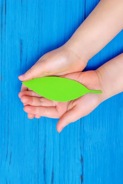 Close up of green paper leaf in hands of child on wooden blue background. Concept of environmental protection. Top view.