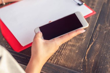 Kafedeki beyaz kağıdın arka planında boş siyah masaüstü ekran ile beyaz cep telefonu tutan bir elin görüntüsü.