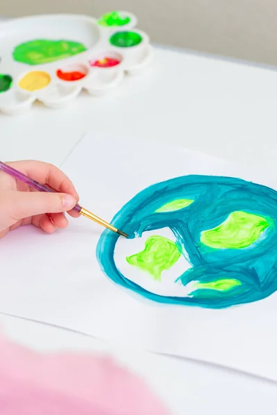 Close up of hand of kid drawing a picture of earth globe. Child is painting Earth with brush by acrylic color on white paper. Earth day concept.