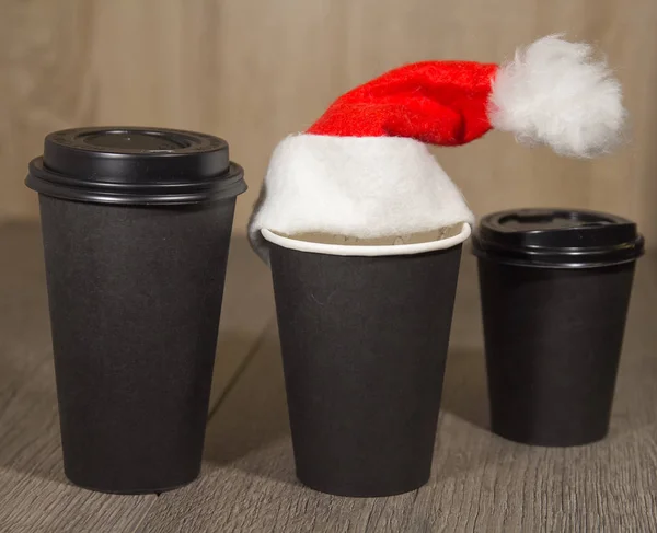 Papel xícara de café preto sobre fundo de madeira, neve em primeiro plano como um acessório ao Natal com chapéu vermelho — Fotografia de Stock