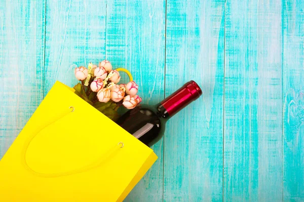 a bottle of wine in a yellow package as a gift, next flowers, gifts, copy space, on blue wooden background, to give girls a set of gentlemen for Dating