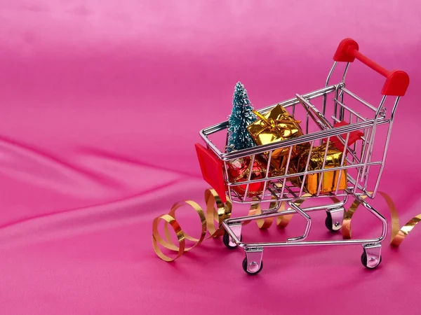 Árbol de Navidad en el carrito de compras en el fondo de satén rosa brillante como fondo, fondo de seda, tarjeta de Navidad. Feliz año nuevo saludos, regalos, símbolos de Navidad en seda rosa — Foto de Stock