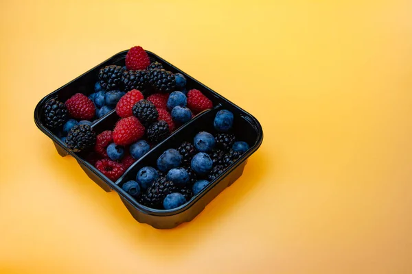 Mix of different fresh berries in black plastic basket sections. Berry selection in basket isolated on yellow background. Sweet berries mix. Raspberrie, blackberry and blueberry isolated.