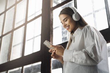 Telsiz kulaklıklı güzel kız akıllı telefondan müzik dinliyor. Mobil aygıtlar için çevrimiçi uygulamalar