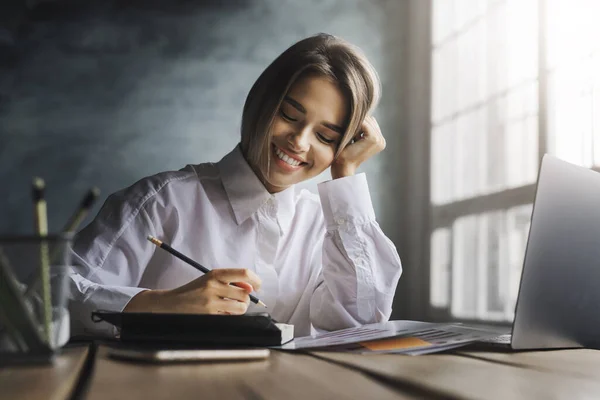 Smiling girl sitting at desk, making notes with pen in copybook. Female student studying in front of laptop