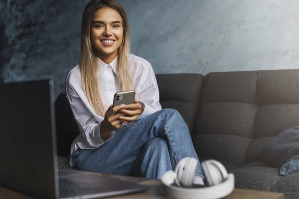 Szczęśliwa uśmiechnięta dziewczyna siedzi na kanapie, trzymając smartfona w dłoni. Młoda kobieta instaluje aplikację messenger do komunikacji — Zdjęcie stockowe