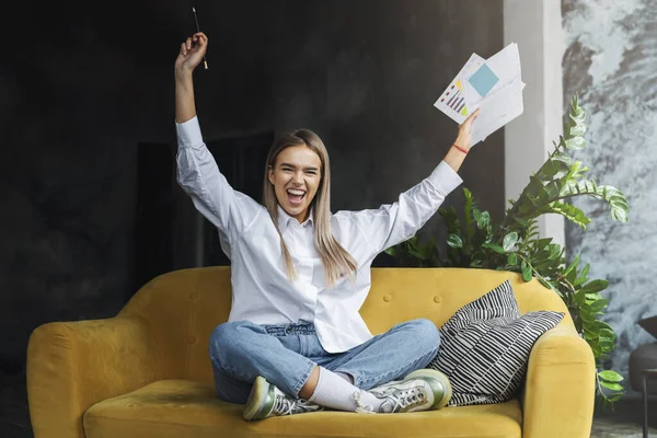 Girl sitting on couch with papers and pen in her hands, being cheerful. Student reacting positively on successful — Stock Photo, Image