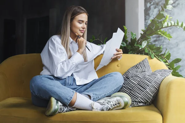 Girl sitting comfortably at home, working with documents distantly. Female reading and making notes with pencil. — Stock Photo, Image