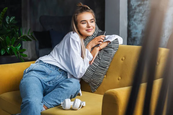 Chica disfrutando de estar en casa, sentado en el sofá, sosteniendo el teléfono inteligente en la mano. Joven hembra usando dispositivo móvil para divertirse — Foto de Stock