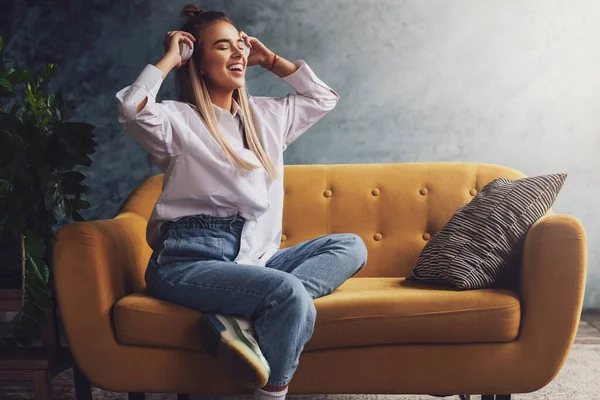 Hermosa mujer rubia en auriculares blancos se sienta en un sofá amarillo, disfruta escuchando podcast. Relajante chica en camisa — Foto de Stock