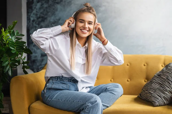 Young beautiful woman listens to music in headphones. Girl in white shirt sits on couch enjoys streaming audio, podcasts — Stock Photo, Image