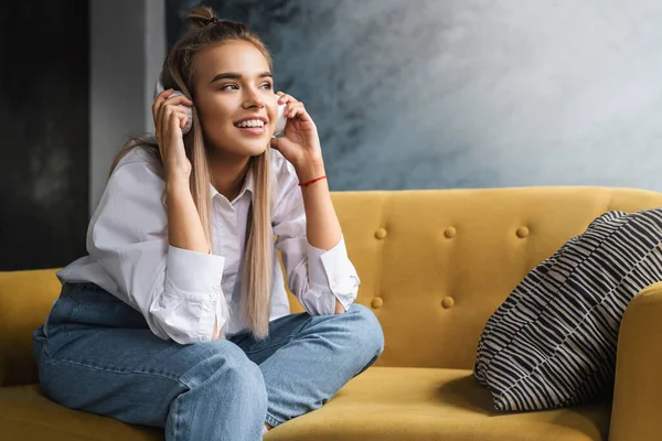 Young beautiful woman listens to music in headphones. Girl in white shirt sits on couch enjoys streaming audio, podcasts — Stock Photo, Image