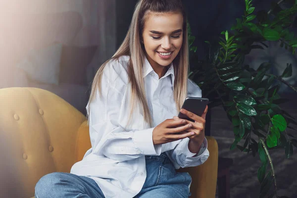 Sorridente bella ragazza bionda siede sul divano giallo e tiene smartphone. Millennial donna comunica con gli amici — Foto Stock
